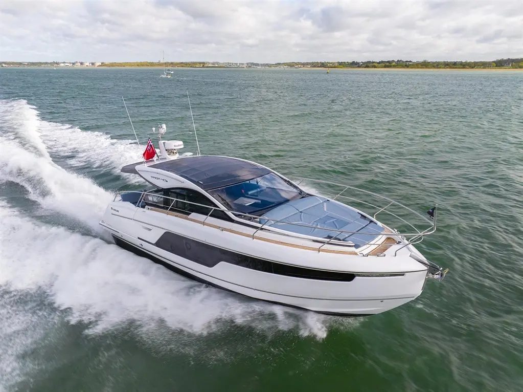 Luxury motor yacht cruising on green water, leaving a wake, with a red flag displayed, under a cloudy sky.